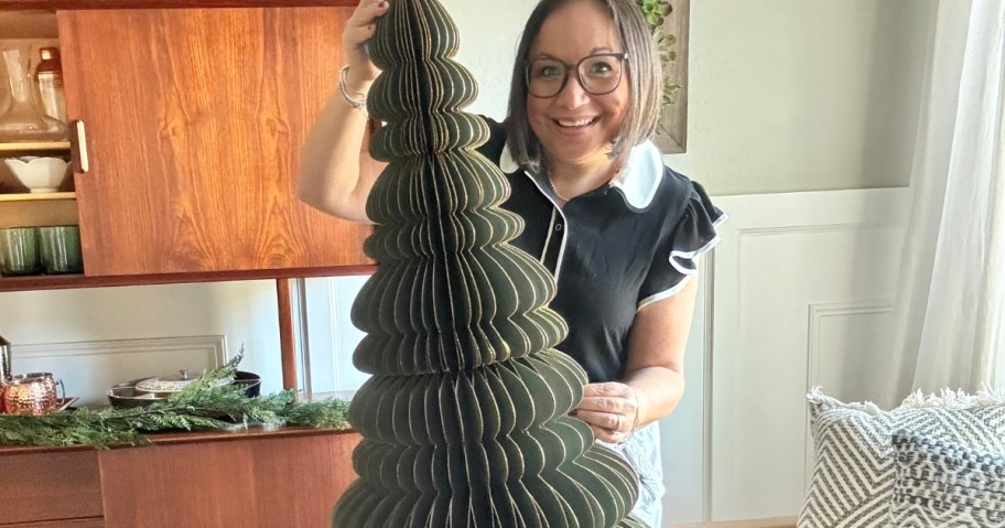 woman with brown hair and glasses smiling, holding up a large green paper Christmas tree