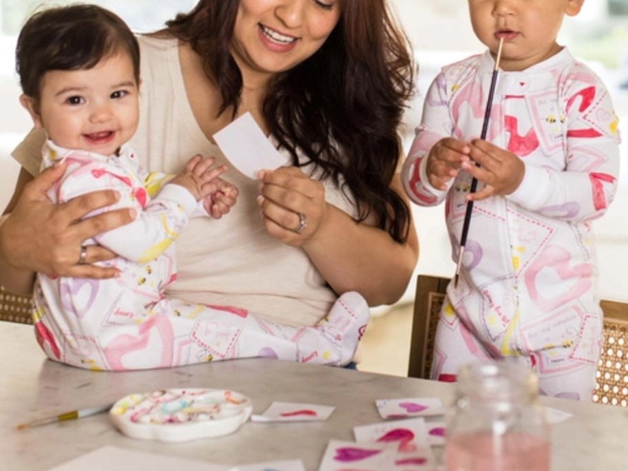 a mom holding a baby and a toddler standing next to them, the baby and toddler are in matching Valentine's themed pajamas