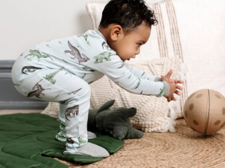 a little boy in a blue footed pajama sleeper that has dinosaurs on it reaching for a ball