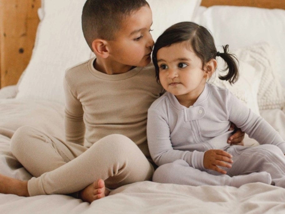 a little boy and younger baby girl in waffle style long sleeve pajamas sitting on a bed
