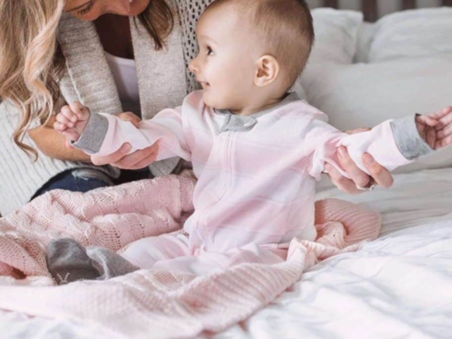 a little girl in a pink ombre footed sleeper sitting on a bed with a woman holding her arms