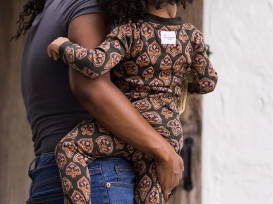 a woman carrying a toddler girl, the toddler is wearing a footed long sleeve pajama in black with pink and orange designs on it