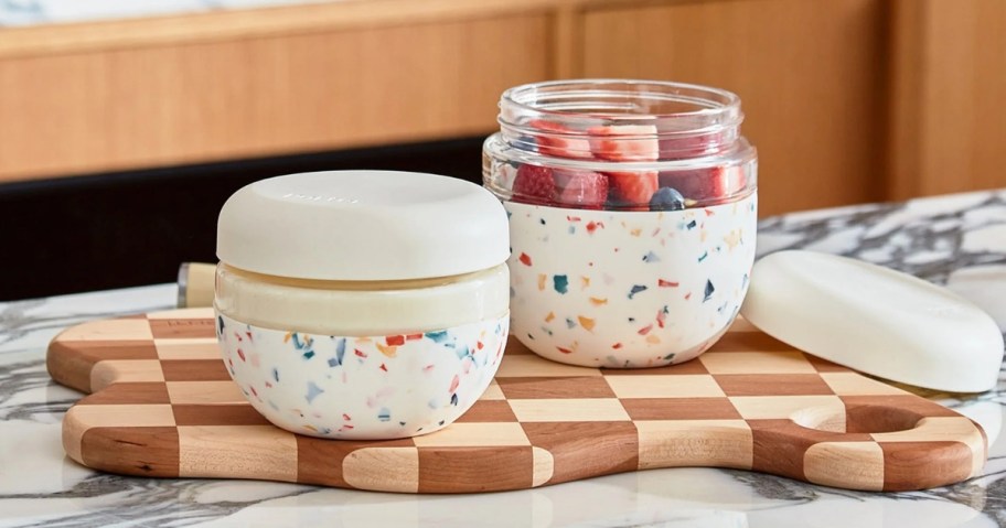 round glass food containers with a white and multi color terrazzo silicone sleeve and white lid sitting on a cutting board on a marble counter, one is open and has strawberries inside