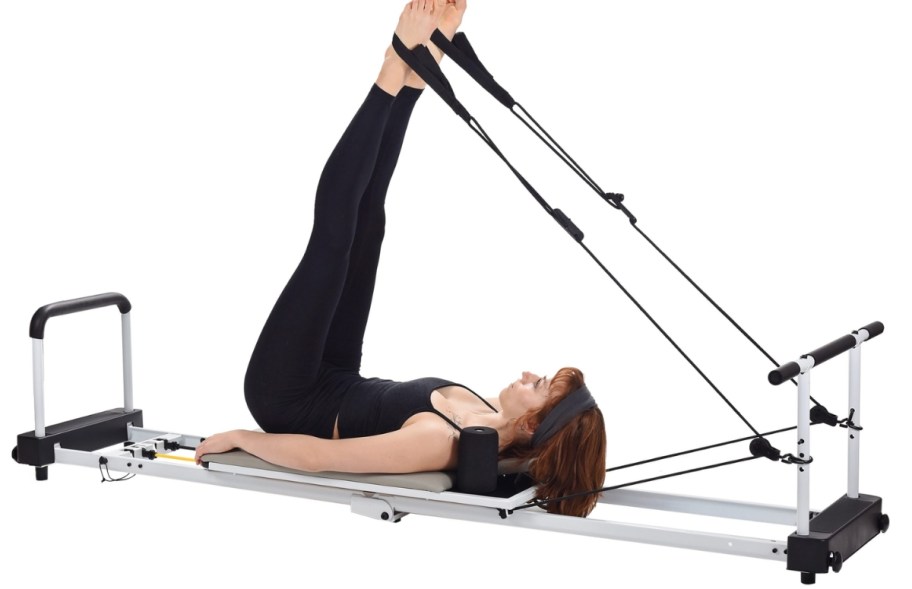 woman using a Pilates machine to workout - her legs are up in the air attached to bands from the machine