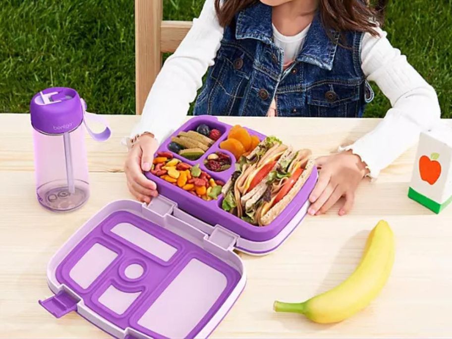Girl eating lunch out of a Bentgo kids lunch box with a Bentgo water bottle next to her