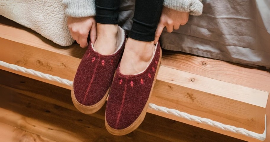 a woman's feet and lower legs propped on a the wood frame of a bed, she's wearing black leggings and wine red clog style slippers that have a small embroidery detail around the opening
