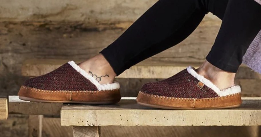 woman's legs sitting on a bench with her feet up on the bench too, she's wearing black leggings and burgundy color clog slippers with a shearling lining