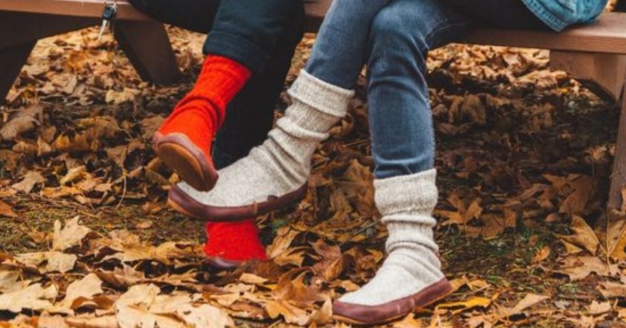 a woman and a man sitting outside at a picnic table, you can only see their legs and they are both wearing jeans and slipper sock shoes, there are leaves all over the ground