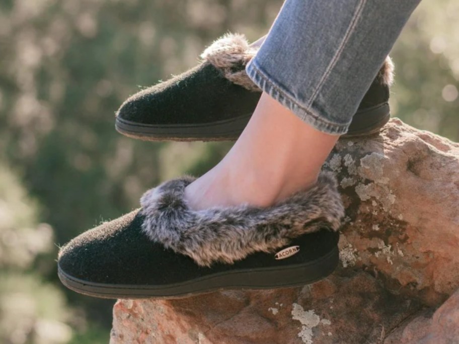 women's feet on a rock, she's wearing jeans and brown slippers with faux fur around the opening of the slippers
