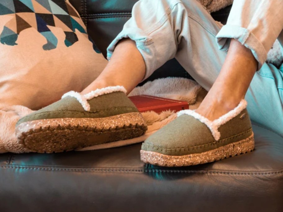 woman's legs, she's sitting on a couch with her feet up on an ottoman and wearing jeans and a olive green slippers with shearling lining