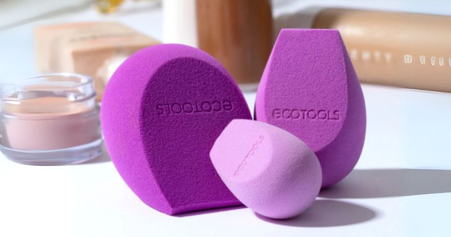 three purple ecotools sponges on table in front of makeup