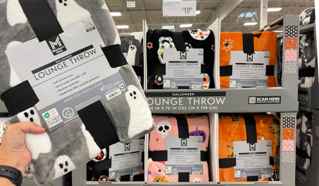 Person holding gray throw blanket with white ghost in front of Halloween blanket display inside Sam's Club store