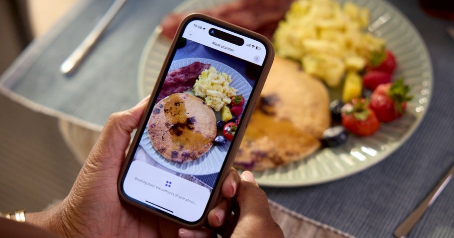 A person uses a phone to scan a breakfast plate with pancakes, scrambled eggs, bacon, and fruit.