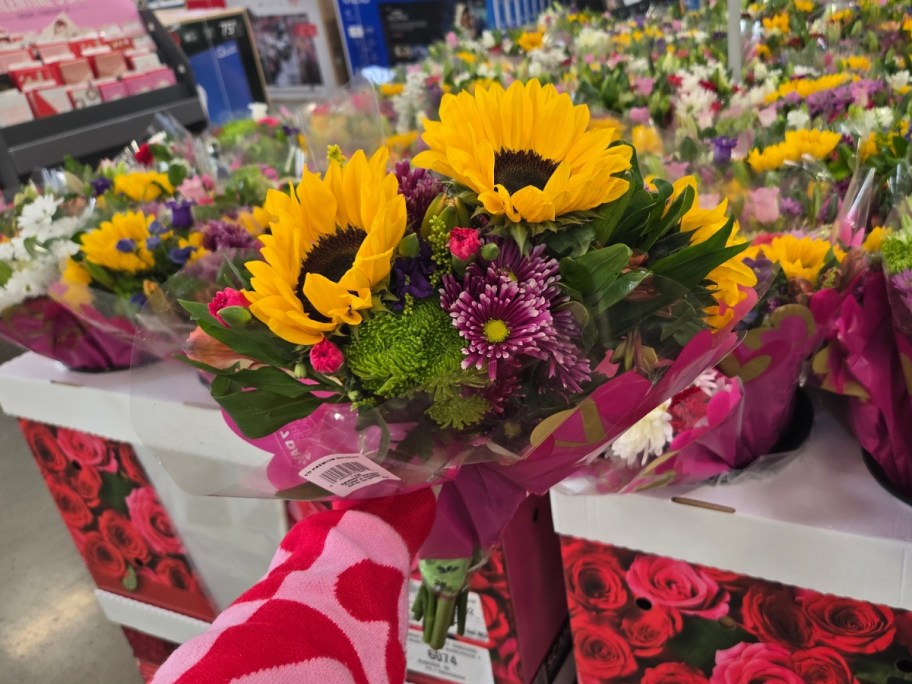 hand holding a bouquet of flowers with sunflowers, and pink and purple flowers wrapped in plastic - a display with more flower bouquets is behind it
