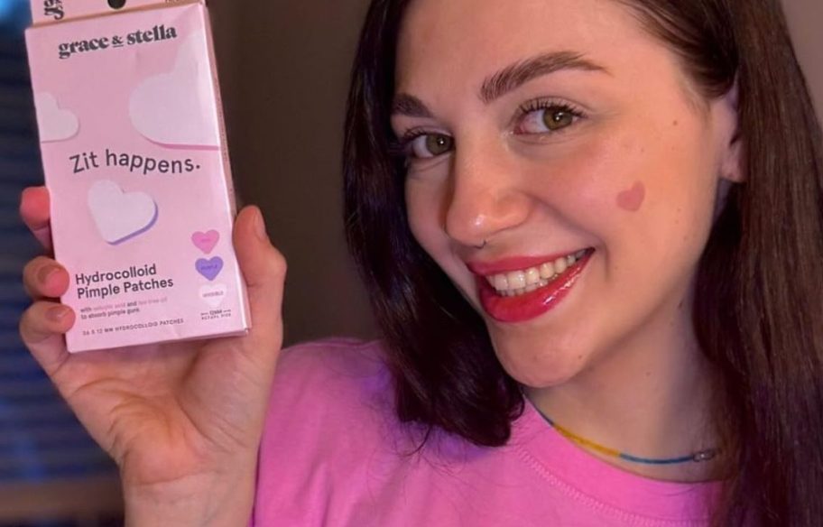 Woman holding a box of Grace & Stella Heart pimple patches while wearing one on her cheek