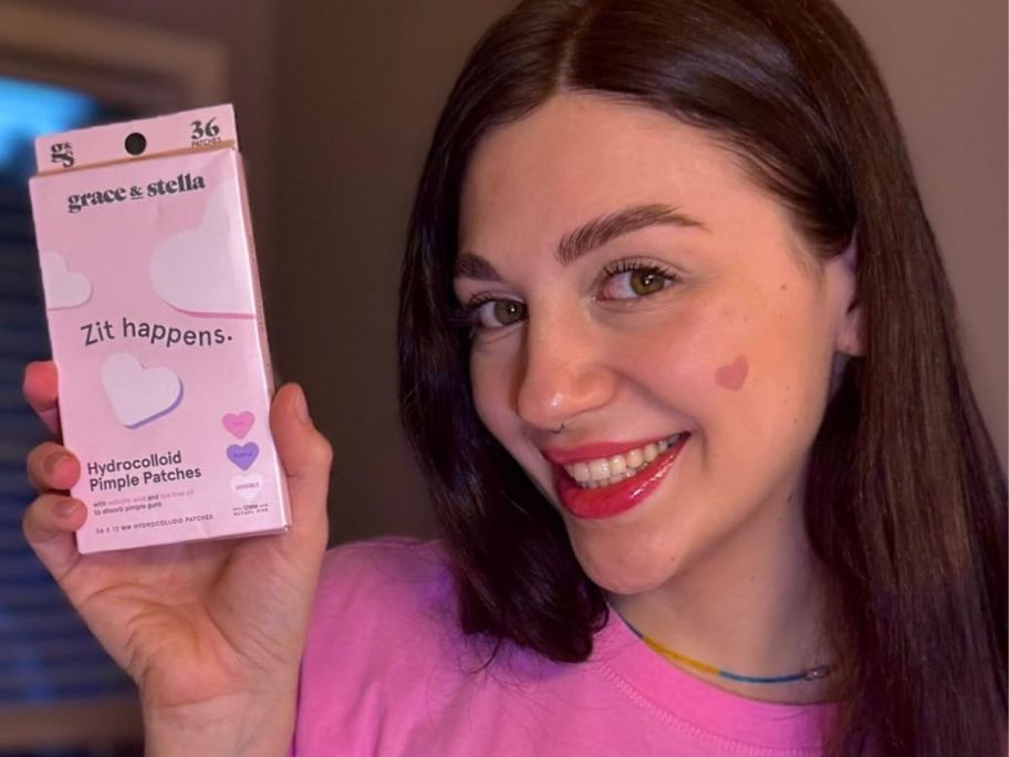 Woman holding a box of Grace & Stella Heart pimple patches while wearing one on her cheek