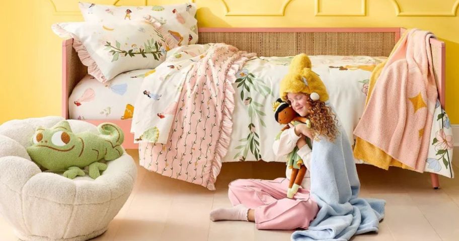 Little girl sitting o floor in front of a bed with Pillowfort Disney Princess Collection Bedding