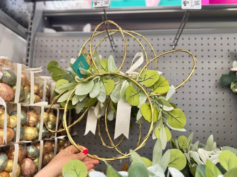 hand reaching for a gold wreath shaped like a bunny rabbit's head that has greenery and flowers on it hanging from a store aisle