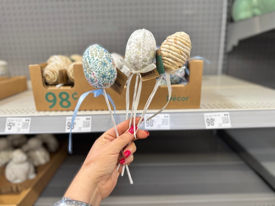 hand holding 3 egg shaped decorative picks with ribbons on them, box with more of them is behind