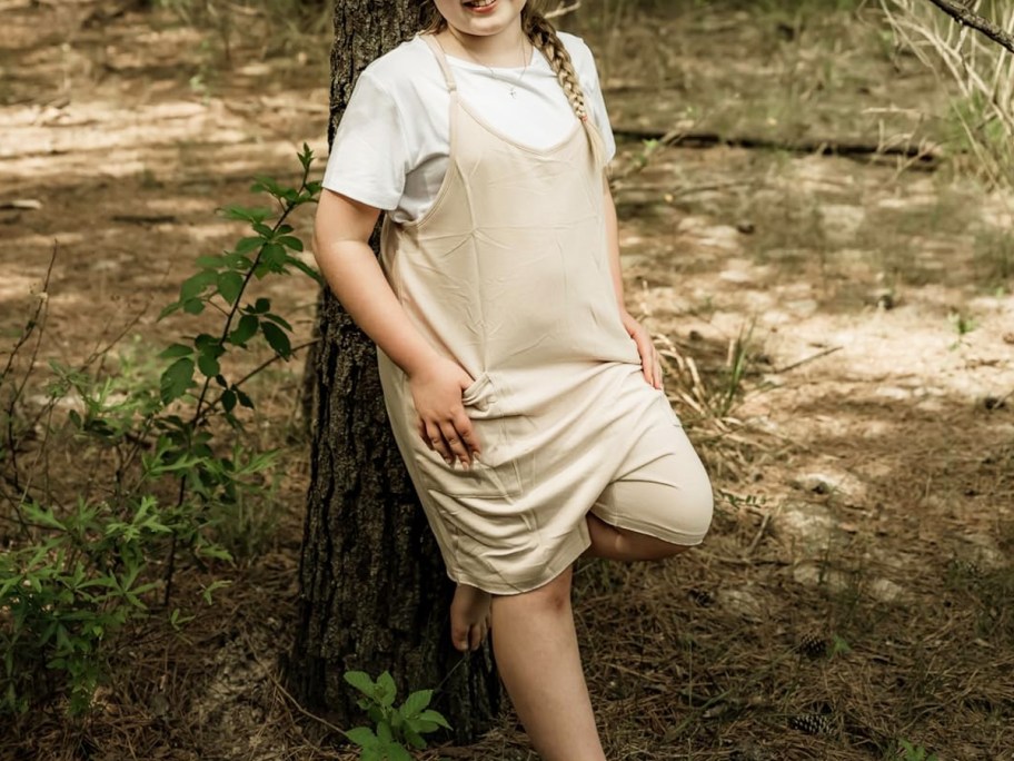 girl in a beige romper with a white t-shirt leaning against a tree