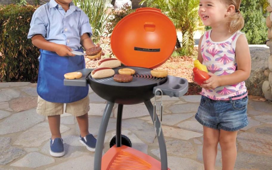 two little kids playing with a pretend grill with toy food and condiments