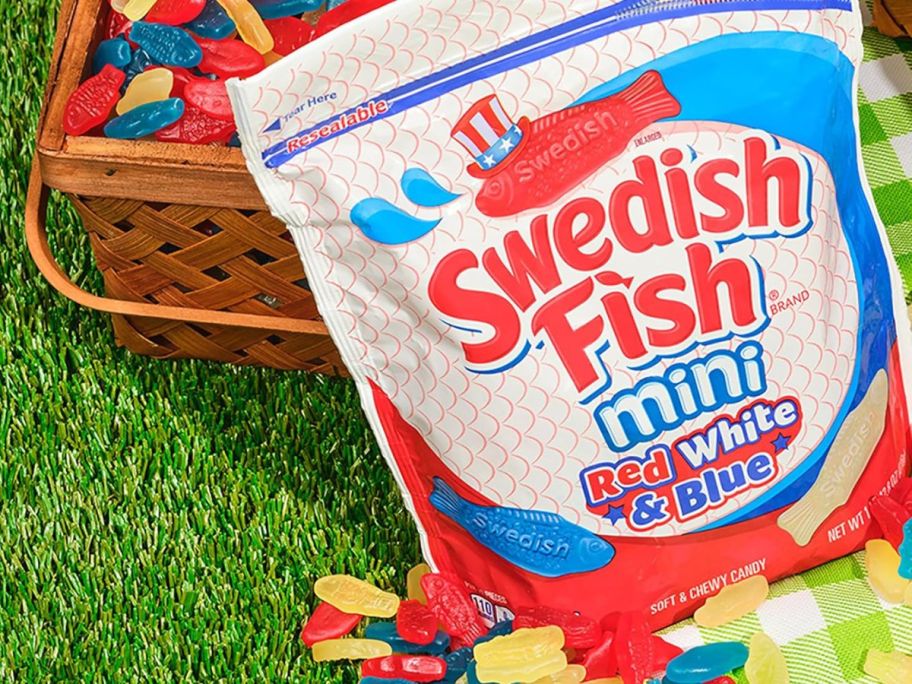 A bag of Swedish Fish on a lawn with a picnic basket