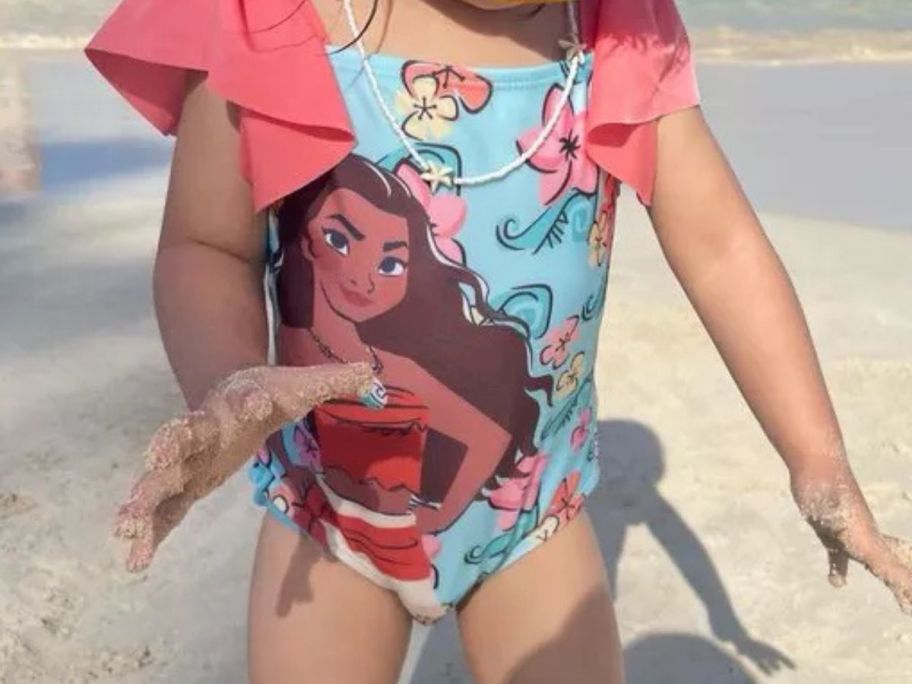 A little girl wearing a Moana swimsuit on a beach 