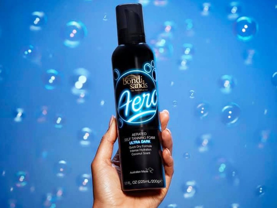 hand holding up a black bottle of Bondi Sands Aero Self Tanning Foam with a blue bubble background