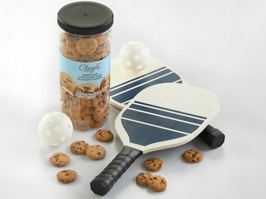 Cheryl's Pickleball gift set featuring two white paddles with navy stripes, two perforated white balls, and a tall container of snack-size chocolate chip cookies scattered around.