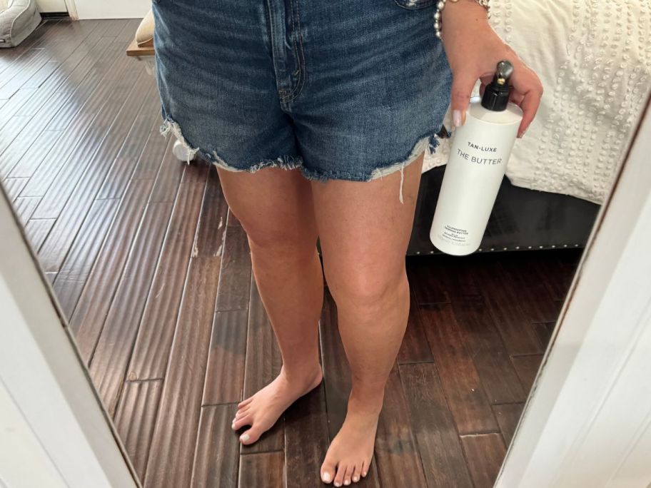 Woman's tan legs in a mirror holding a big bottle of Tan Luxe The Butter Lotion