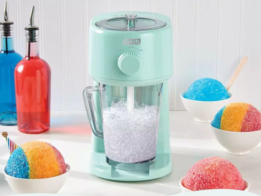 A Dash Deluxe Shaved Ice & Slushy Maker surrounded by syrups and treats