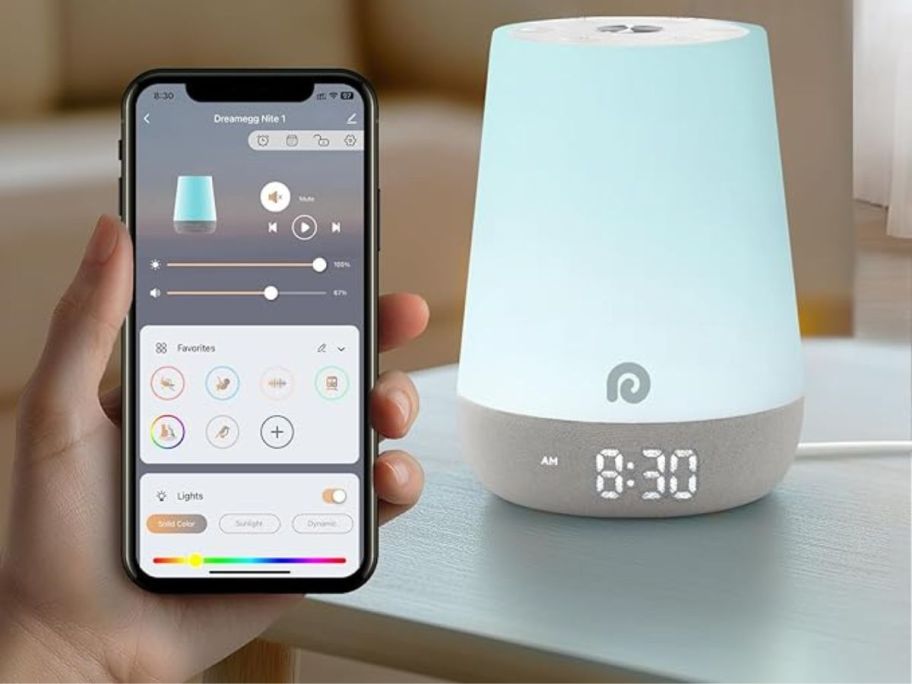 A Dreamegg Sound Machine on a table emitting a light blue light next to a person with a phone