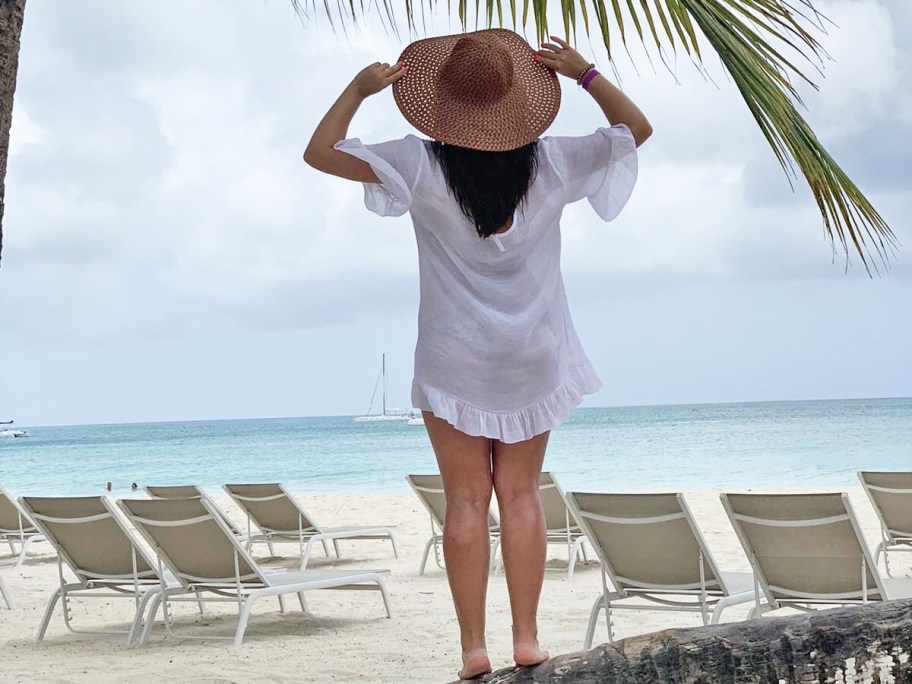 woman standing on tree on beach in a white swimsuit coverup and a sun hat