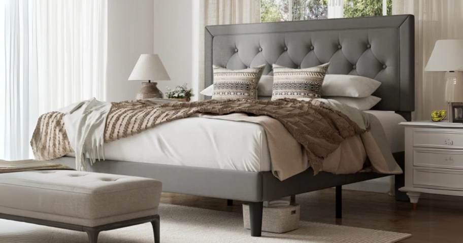 a bedroom with a bed in a grey upholstered bed frame, fabric bench and nightstand