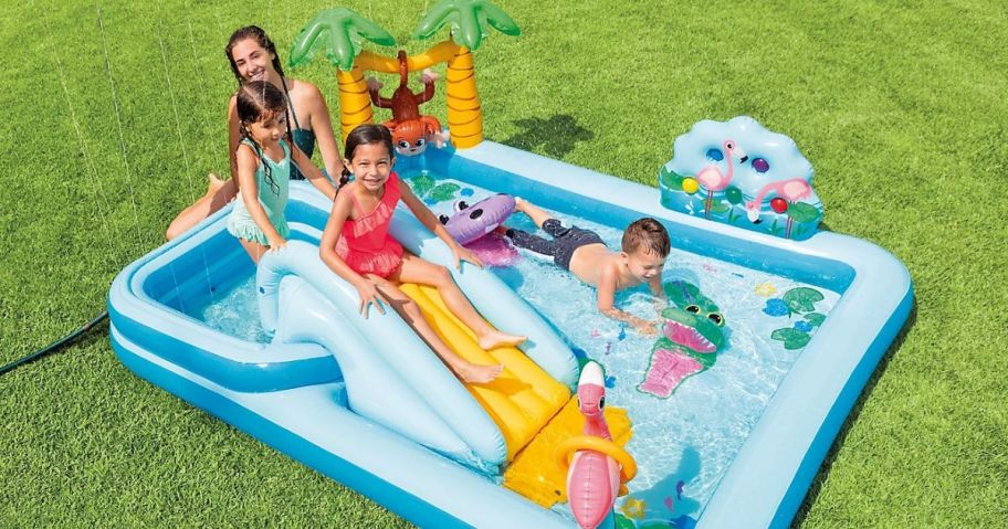 Kids and a woman are playing in an inflatable pool for kids with sprinklers. 