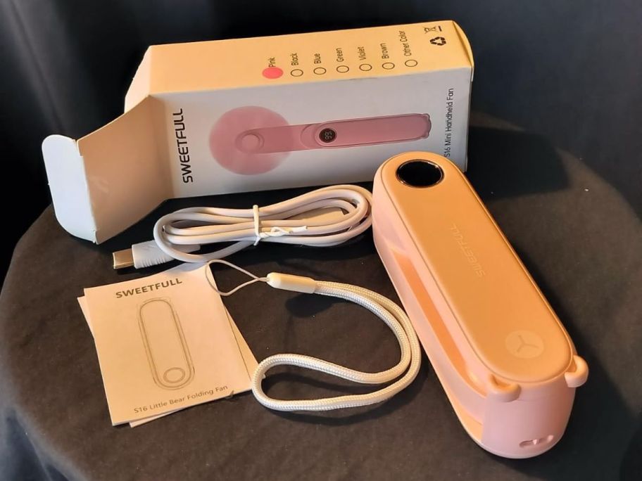 A small, pink fan with charging cable, wrist fob, and instructions