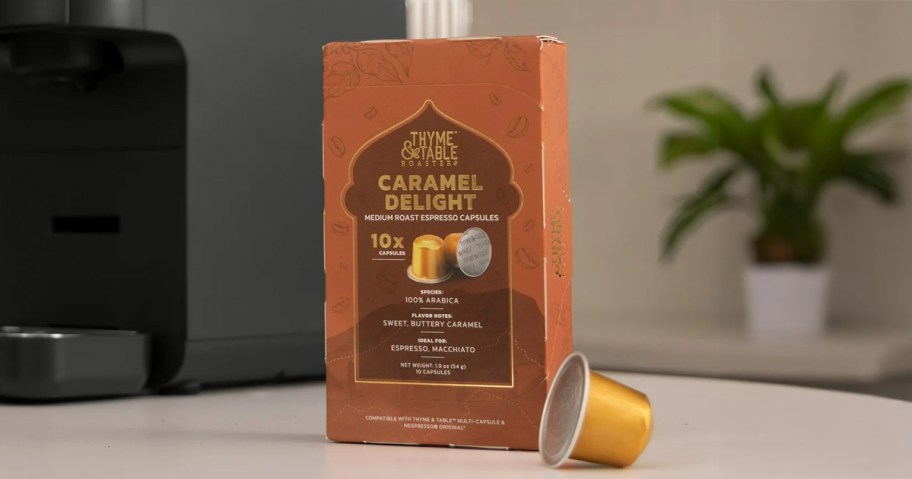 Box of Thyme & Table Caramel Delight medium roast espresso capsules with one gold capsule in front, positioned beside a coffee machine on a kitchen counter.