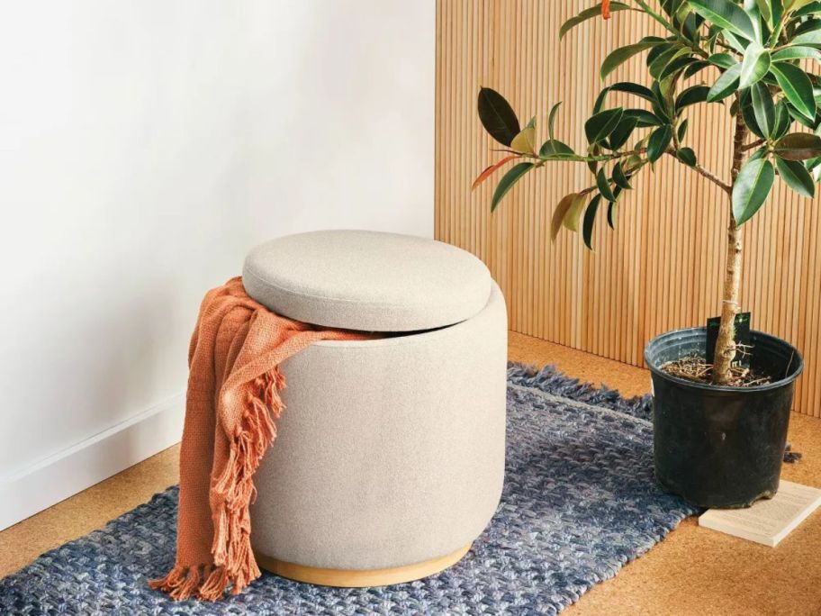 cream colored Room Essentials Round Upholstered Storage Ottoman with blanket sticking out of it on floor