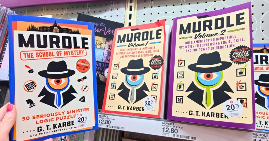 hand holding a Murdle mystery puzzle book with more on a store shelf behind it