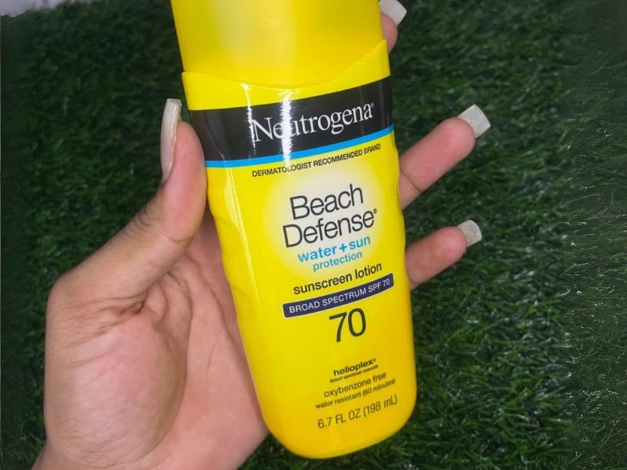 hand holding Neutrogena Sunscreen Lotion Beach Defense SPF 70 bottle above grass