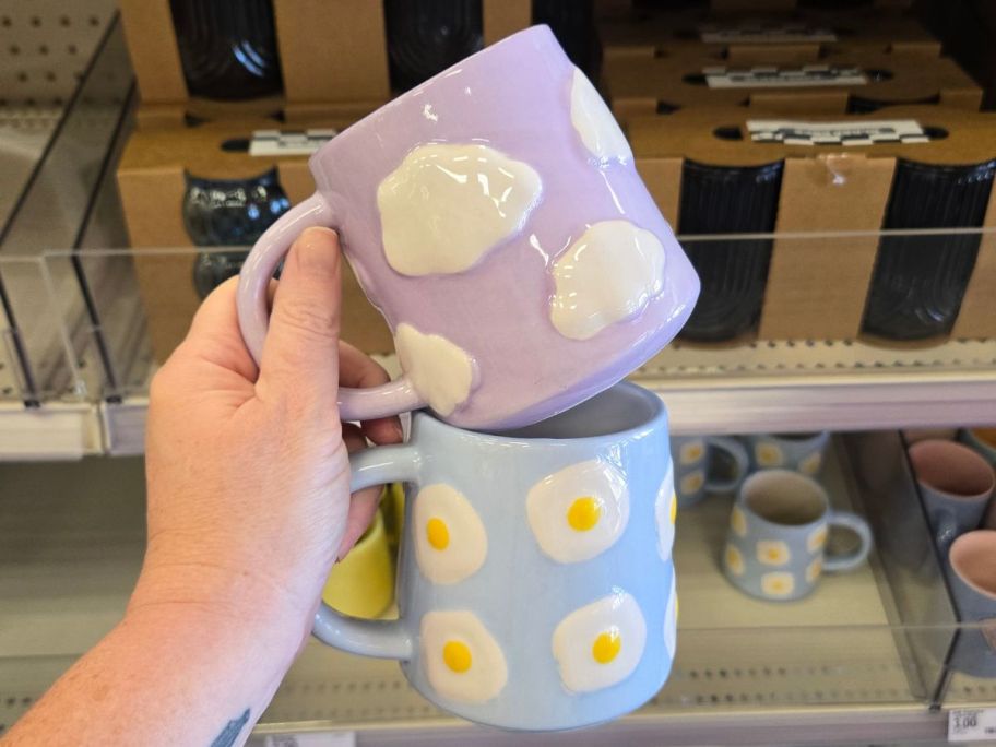 Hand holding two coffee mugs with one purple with clouds and one blue with fried eggs