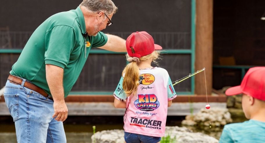 Cabelas fishing instructor helping a girl wearing a cabelas hat and t-shirt fish in their lake