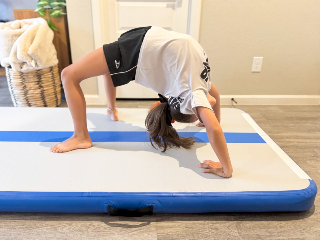 Young girl doing backbend on inflatable blue and white gymnastics mat inside home