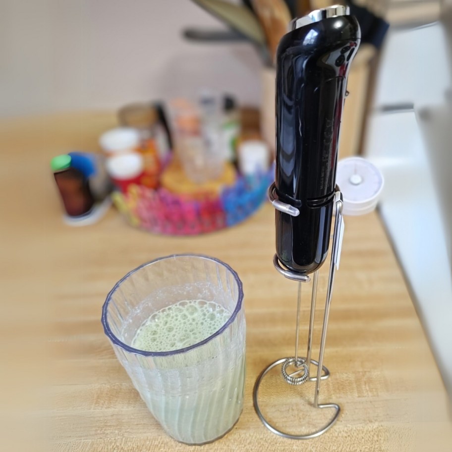 a milk frother on a stand next to a cup of frothed milk on a kitchen counter