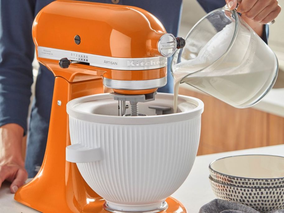 Ice cream maker attachment being used on an orange kitchen aid with hand pouring the mixture inside