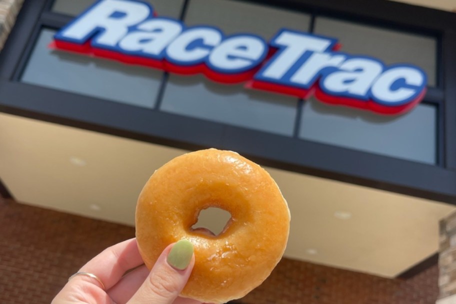 Hand holding a single glazed doughnut, outside in front of the RaceTrac store, in front of their store sign.