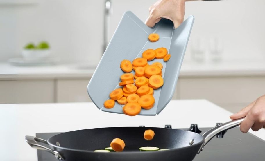 a womans hand holding a foldable cutting board as she dumps chopped carrots into a saute pan.