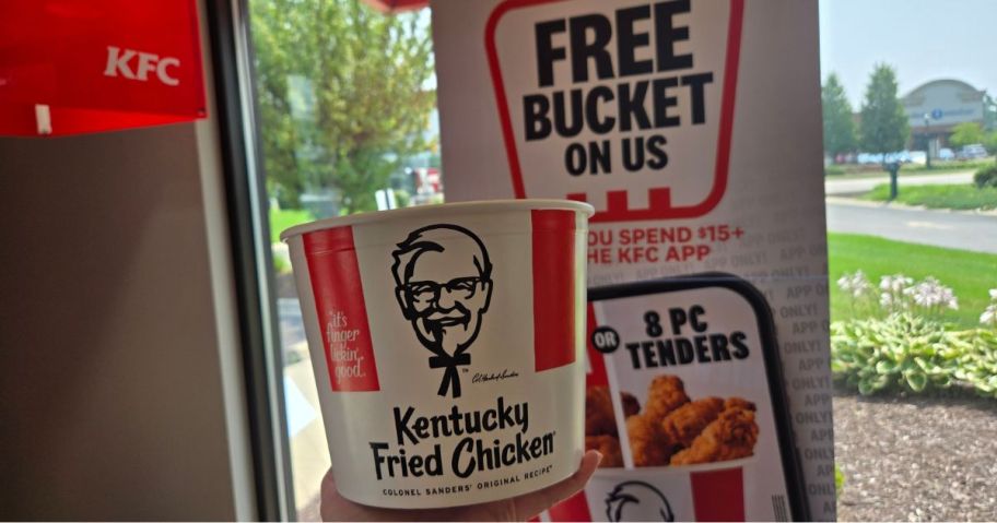 hand holding bucket of kfc chicken in front of "free bucket on us" sign in store