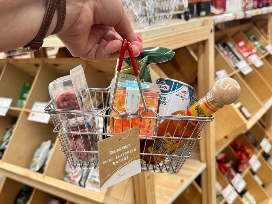 a womans hand holding a mini shopping basket filled with snacks