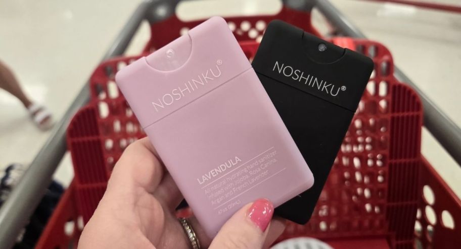 hand holding two mini hand sanitizers in pink and black in front of a target shopping cart